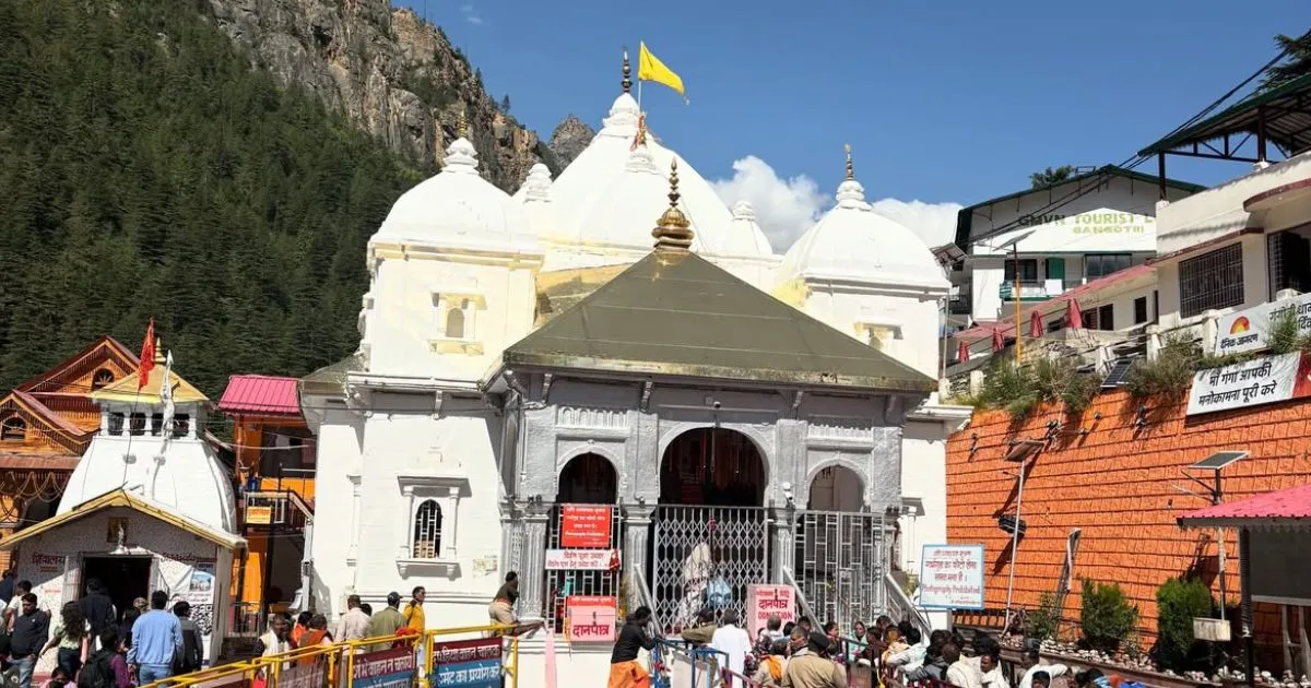 Char Dham Yatra Registration, Route Map, Itinerary, Packages & Helicopter Booking 7 A beautiful white temple structure featuring a yellow flag on its dome stands near a forested hill as visitors line up at the entrance.