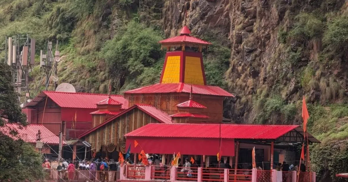 Char Dham Yatra Registration, Route Map, Itinerary, Packages & Helicopter Booking 6 A striking temple structure with distinct red roofing built closely against a steep, rocky cliffside as crowd of devotees gather nearby.