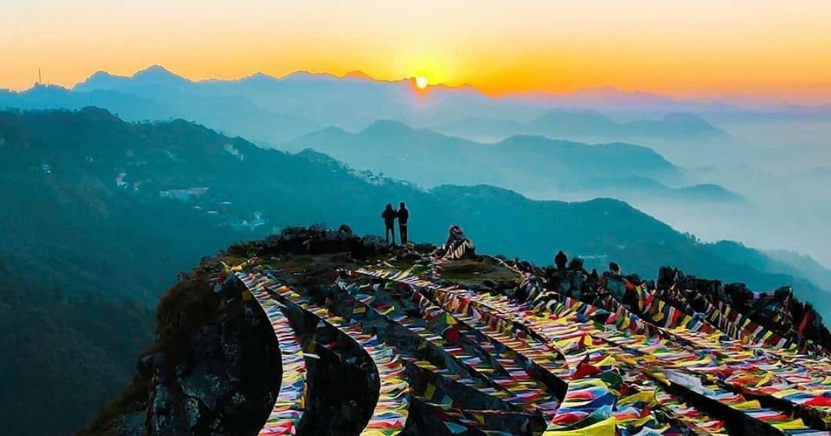 Two hikers enjoy a vibrant sunset over mountains, adorned with colorful prayer flags. A beautiful trek and one of the Easy Treks Near Dehradun.