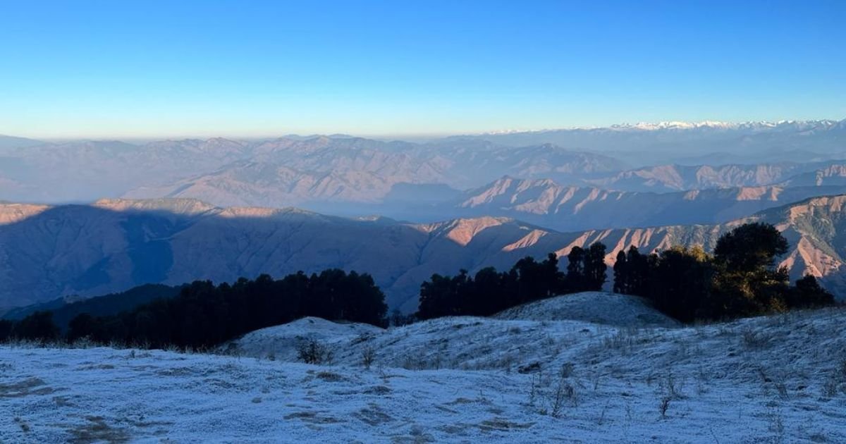 Frost-covered ground and scattered trees offer panoramic views of distant, hazy mountains. Discover stunning vistas on Easy Treks Near Dehradun.