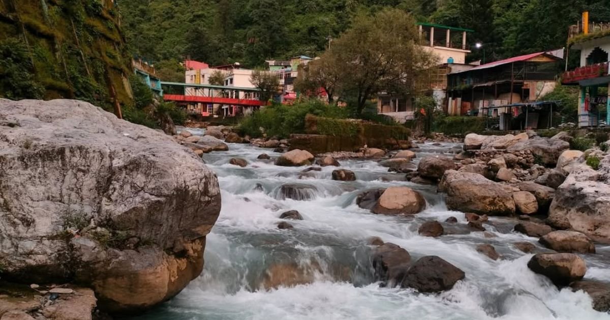 A strong river flows over large rocks with some buildings in the background. Enjoy the calming river on Easy Treks Near Dehradun.
