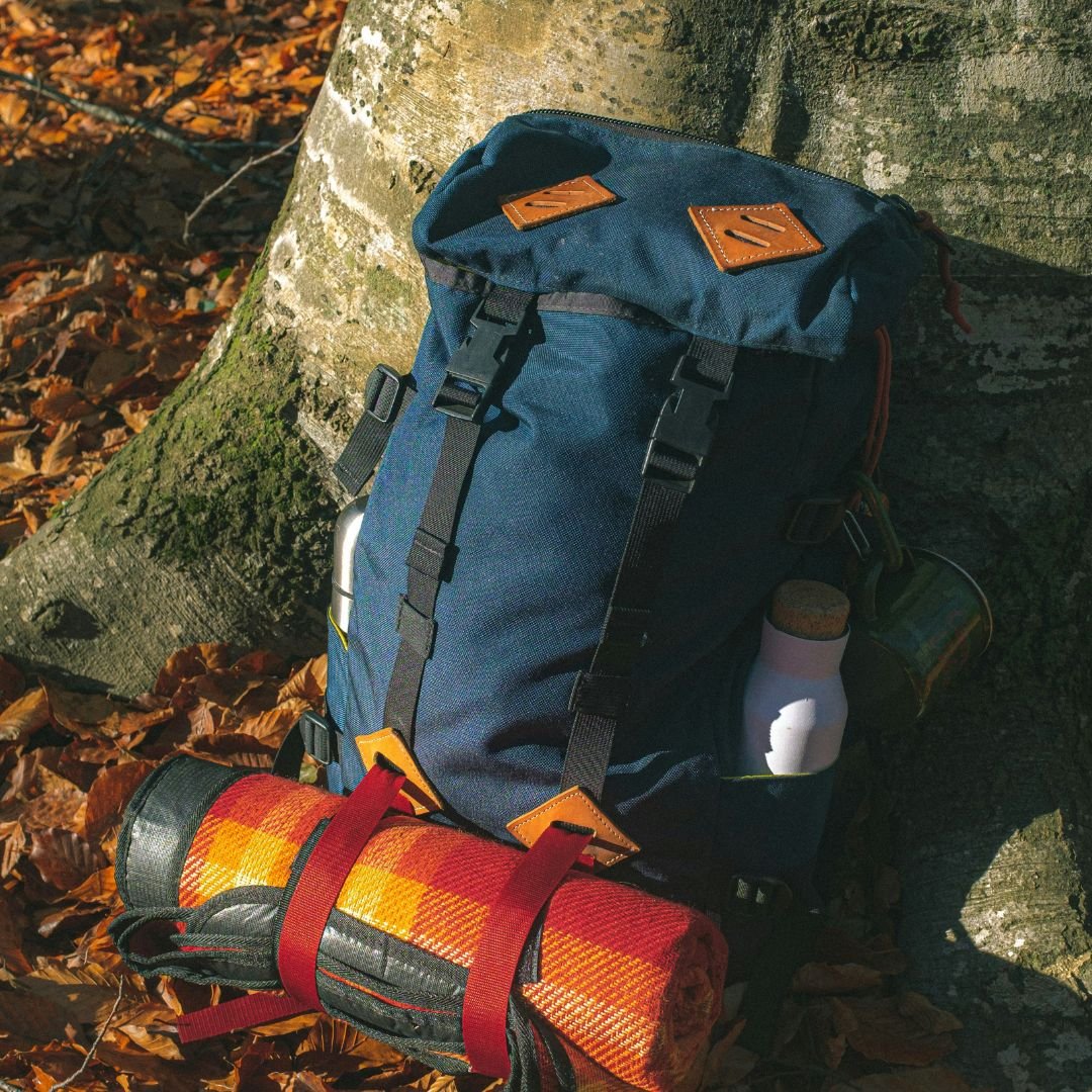 A blue backpack and a rolled-up blanket are leaning against a tree trunk, ready for a Kedarnath itinerary. This is what you should pack.